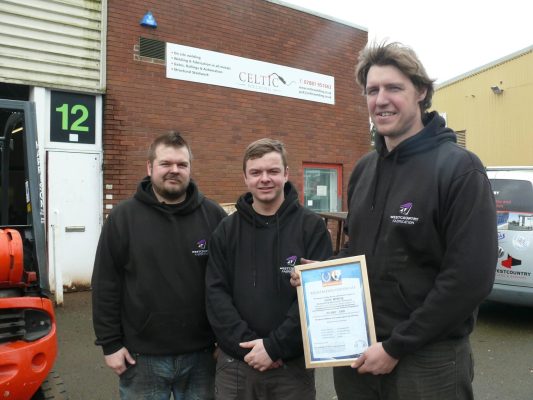 Three workers pose with their first ISO9001 certificate outside of a workshop in Plymouth