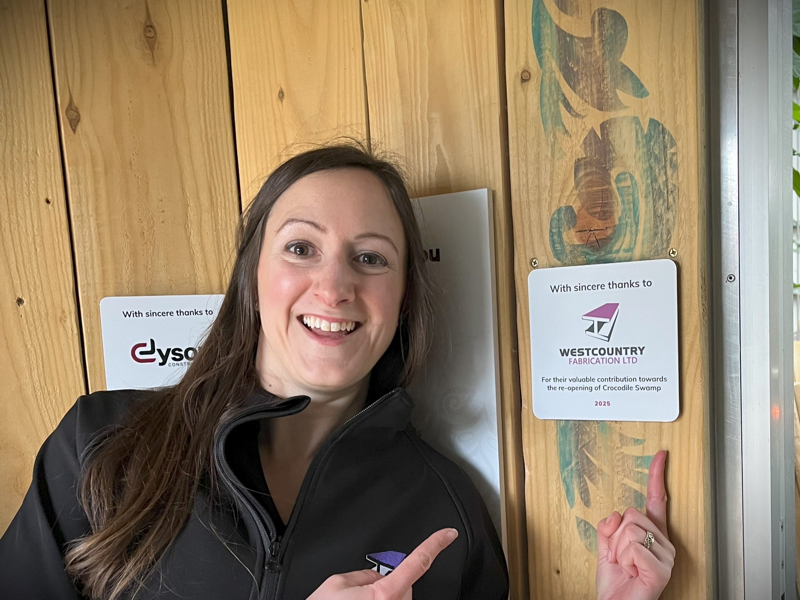 Person smiling pointing to a sign that says 'With sincere thanks to Westcountry Fabrication Ltd For their valuable contribution towards the re-opening of Crocodile Swamp 2025'